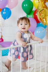 Fototapeta na wymiar Little girl with red hair and dark eyes,wearing a gray dress with white and pink polka dots,standing on a white bed in the background of colorful balloons and gold stars on his first birthday