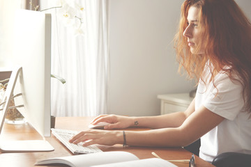 Cropped portrait of young confident woman entrepreneur typing on the computer, female freelancer with red hair and in white T-shirt keyboarding on the PC sitting against home interior background
