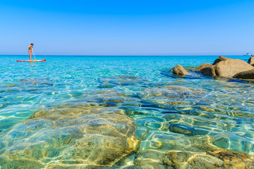 Crystal clear sea water of Bodri beach and unidentified woman swimming on surfboard in distance, Corsica island, France