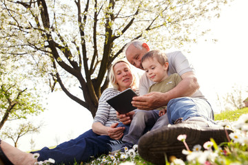 Fototapeta premium Grandparents with grandson enjoying the sunny spring day outdoors. They are looking something on tablet.