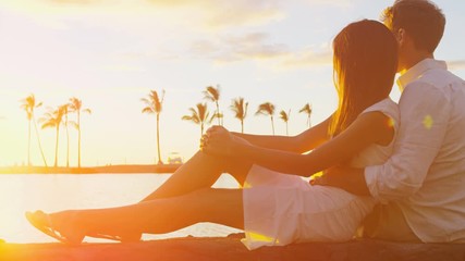 Couple romantic sunset happy in love embracing enjoying travel vacation at beach sitting down looking at ocean view in embrace on honeymoon holidays getaway. Interracial couple. RED EPIC. - Powered by Adobe