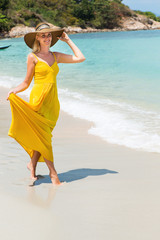 Carefree beautiful elegant blonde woman in beach straw hat holding her long yellow dress and walking barefoot by sand of tropical oceanshore. Natural woman beauty. Lady dancing near water and smiles.