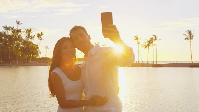 Selfie couple romantic at sunset on vacation travel taking photo in love at beach using smart phone holding around each other in embrace on honeymoon holidays getaway. Man and woman. RED EPIC.