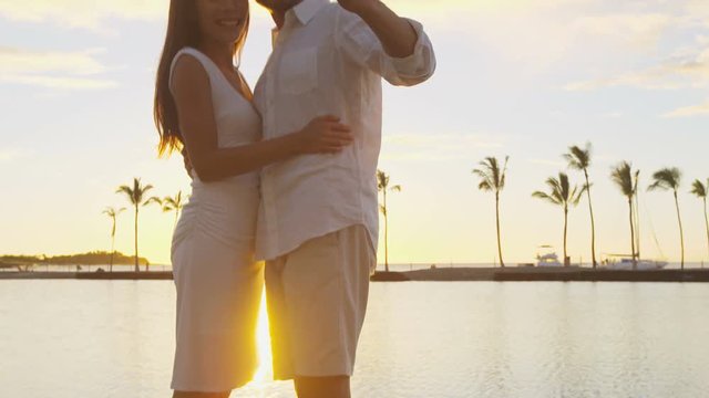 Kissing couple romantic at sunset on vacation taking selfie in love at beach using smart phone holding after kiss and embrace on honeymoon holidays getaway. Man and woman. RED EPIC.