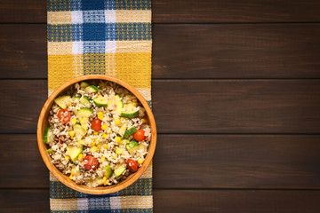 Rice dish with mincemeat and vegetables (sweet corn, cherry tomato, zucchini, onion) in wooden bowl, photographed on dish towel on dark wood with natural light