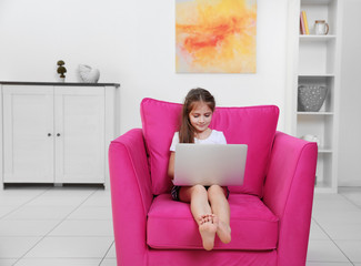 Fototapeta premium Little girl using laptop while sitting in a pink armchair.