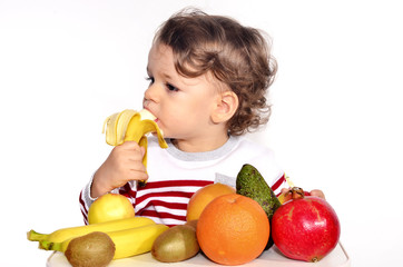 Fototapeta premium Kid having a table full of organic food. Cheerful toddler eating healthy salad and fruits. Baby choosing between apples, bananas, oranges, avocado,pomegranate. kiwi. Isolated on white