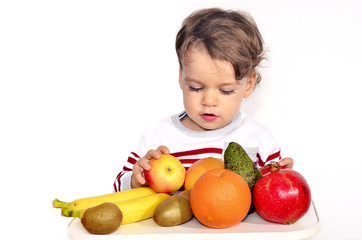 Fototapeta premium Kid having a table full of organic food. Cheerful toddler eating healthy salad and fruits. Baby choosing between apples, bananas, oranges, avocado,pomegranate. kiwi. Isolated on white
