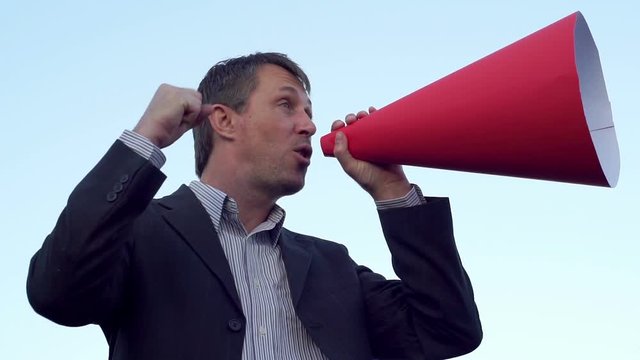 Side view shot of a man wearing a suit standing against a clear blue sky and making an announcement through a red rolled paper bullhorn while shaking his fist and pointing his finger.