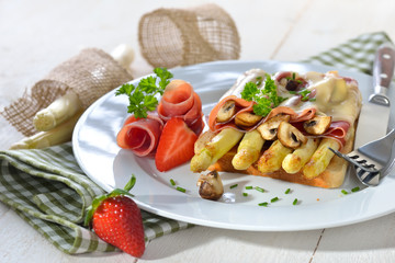 Überbackener Toast mit gebratenem weißen Spargel, Serrano-Schinken, Champignons und einer Scheibe Schnittkäse, mit Erdbeeren serviert -  Baked toast with fried asparagus, ham, mushrooms and cheese