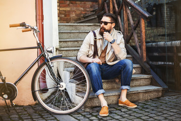 Obraz na płótnie Canvas stylish hipster bearded man in the street bike vintage leather handbag, glasses, smoking pipe. Vintage style photography, lighting effects. Old european street city