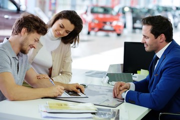 Fototapeta premium Happy young couple signing a contract to purchase a new car at the dealership showroom.