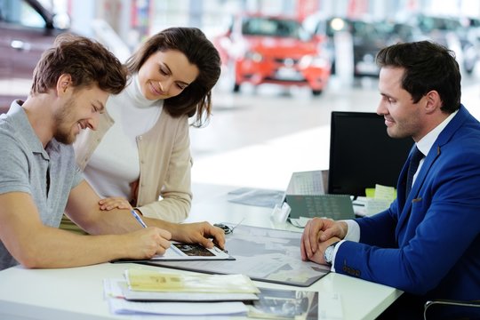 Happy young couple signing a contract to purchase a new car at the dealership showroom.