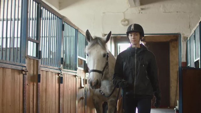 Young jockey girl is walking with white horse in a stable. Shot on RED Cinema Camera.