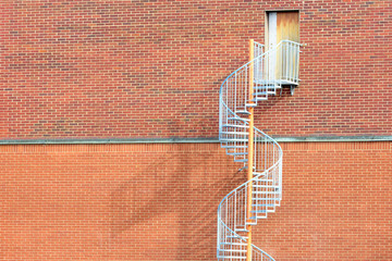Spiraling metal stairs up a red brick wall to a door without handle. This is an emergency exit, not meant to be entered from the outside. Copy space.