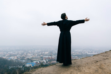 Priest bearded man raised his hands up on the hill above the town. Pastor posing outdoors in...