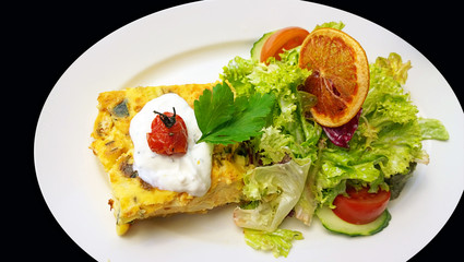 Quiche and salad topped by an orange slice, white saucer isolate on black