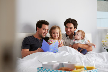 Male gay parents relax in bed with kids, food in foreground