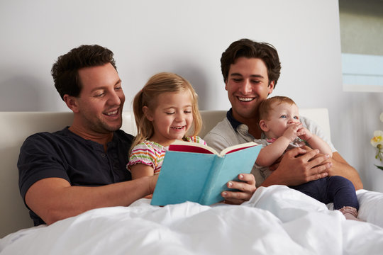 Male Gay Parents Reading A Book In Bed With Two Kids