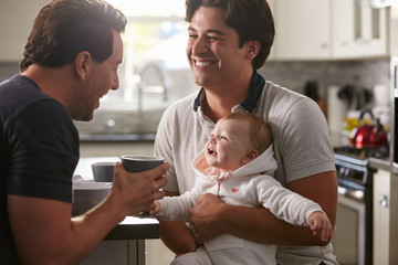 Male gay couple holding baby girl in their kitchen - obrazy, fototapety, plakaty
