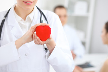 Female doctor with stethoscope holding heart.  Doctor and patient sitting in the background