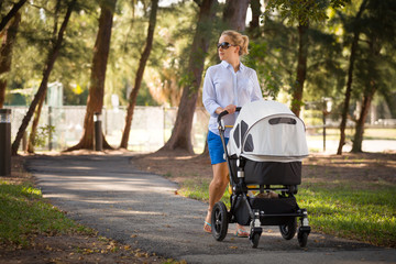 Fototapeta na wymiar Young mother walking and pushing a stroller in the park. Mother strolling with newborn. Beautiful happy mother with pram outdoors. Summer walks in the sunny day. Girl with carriage.