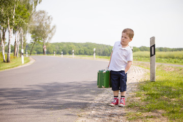 Adorable little kid boy in a white polo shirt and shorts holds a green retro suitcase and  stands on the countryside road waiting for a bus or a car. Ready for travel. Vacation time. Child on the road