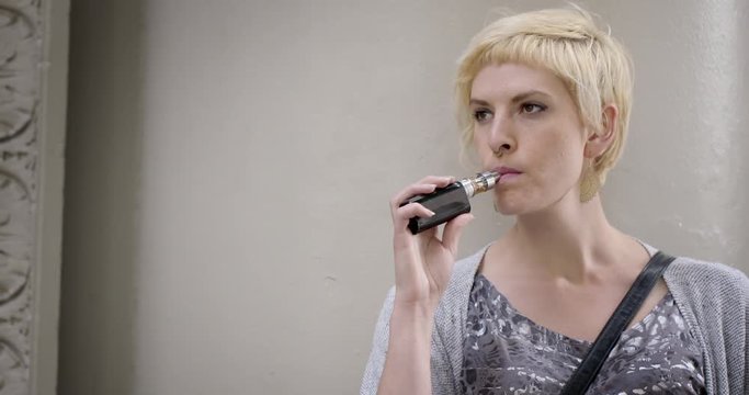 Medium close up of woman using an electronic cigarette in an urban setting.  Hand-held camera, profile view recorded at 60fps.