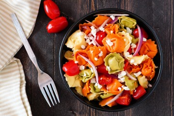 Bowl of colorful tortellini pasta salad with tomatoes and onions, overhead scene on dark wood table