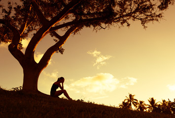 Sad young woman sitting outdoors under a tree.