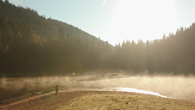 Countryside couple  in ukrainian ethnic clothes tenderly embracing on the rocky shore on sunset in golden mist at the picturesque mountain lake in Carpathians. Eternal harmony between human and nature
