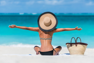Summer vacation happy carefree joyful bikini woman arms outstretched in happiness enjoying tropical beach destination. Holiday girl sitting with sun hat relaxing from behind on Caribbean vacation.