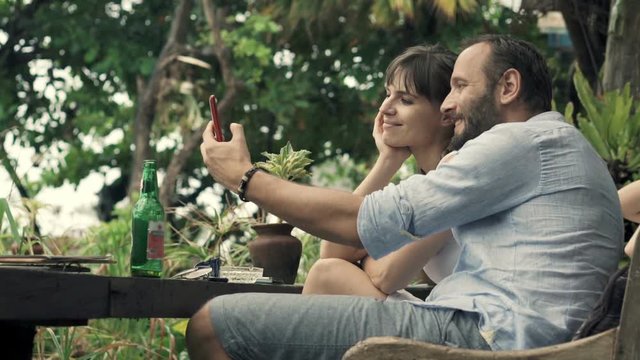 Happy couple taking selfie photo with cellphone in outdoor cafe in garden
