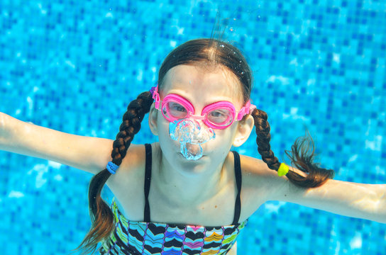 Child swims in pool underwater, funny happy girl in goggles has fun under water and makes bubbles, kid sport on family vacation

