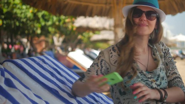 Pretty girl on a beach taking selfie with hir smartphone. Beautiful woman in a straw hat and sunglasses on a lounger. Tropical vacation in Vietnam.