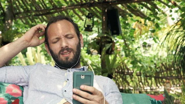 Sad, unhappy man listen music on cellphone sitting on sofa in garden 
