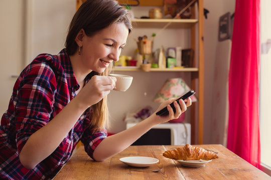 Young woman sitting at a table drinking tea at home, smiling and looking at your phone. On the table there is a croissant.