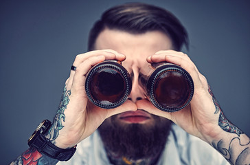 Bearded hipster man holding beer bottles like binocular.