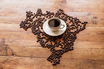 A cup of coffee standing on coffee beans laid out in the shape of Brazil, on a wooden table