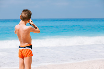 Beach vacation dream. Handsome young boy enjoying in beautiful tropical beach and taking some photos with his camera.