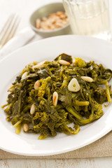 boiled greens with nuts and garlic on white plate and glass of wine on white wooden background