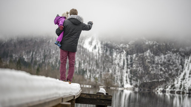 Young father standing on the end of a snowy pier in a lake holdi