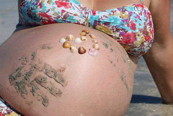Belly in ninth month of pregnancy on the beach with sand handprints and heart of shells on it