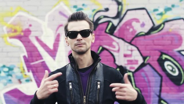 Young man in sunglasses gesture in camera. Read rap. Graffiti colorful wall on background. Subculture. Artist