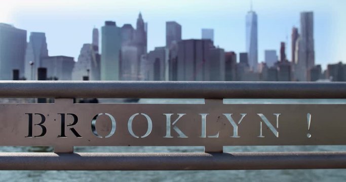 Iconic view of a metal railing south of the Brooklyn Bridge reading "BROOKLYN!" The Financial District is visible in the background as Ferryboat sails into the frame. 