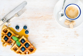 Espresso coffee cup, soft belgian waffles with fresh blueberries and marple syrup on white painted wooden board over light blue background