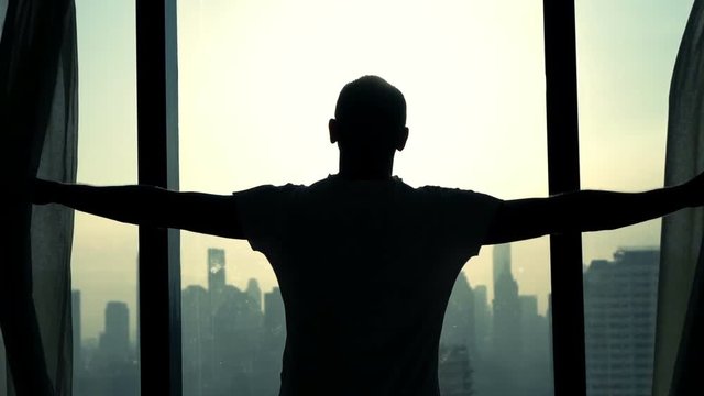 Young man unveil curtains and admire cityscape view, slow motion
