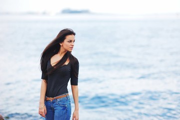 beautiful brunette girl in blue jeans and a black blouse standing on background of water