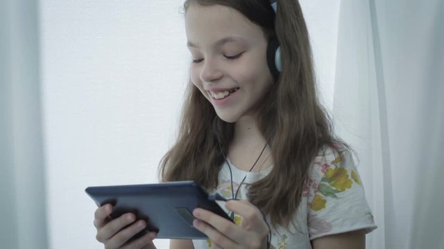 Beautiful little girl with headphones watching funny videos on tablet and laughs.