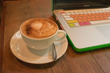 Hot coffee in white cup and saucer with silver spoon on wooden table with laptop and colorful keyboard and case in day time/ Hot coffee in white cup and saucer